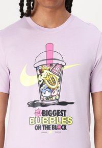 Purpura T-krekls ar karikatūru stila dzēriena kausa grafiku, ar krāsainiem elementiem un tekstu "NAKAMIE LIELĀKIE BURBUĻI BLOKĀ".