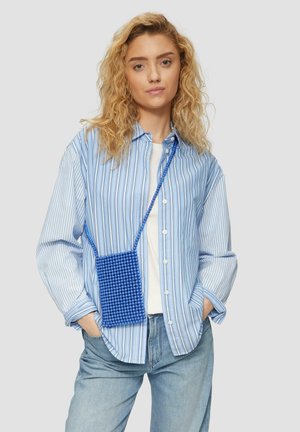 Chemise à rayures bleues et blanches à manches longues, portée par-dessus un t-shirt blanc, avec un sac bandoulière rectangulaire en perles bleues.