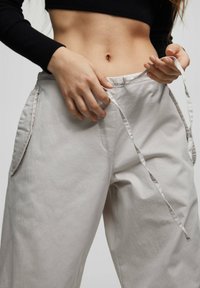 Kvinna klädd i ljusbeige byxor med knytband i midjan och en svart kortärmad topp, hennes händer justerar byxornas knytband.