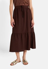 Falda marrón, de longitud media y con capas, hecha de tela texturizada con cintura ajustable. Combinada con sandalias de tono dorado.