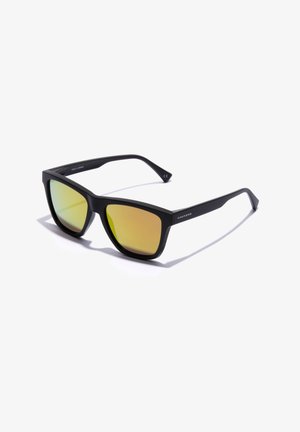 Gafas de sol de plástico negro con un diseño angular y lentes tintados en verde. Las varillas presentan un detalle de marca simple, fabricadas en España.
