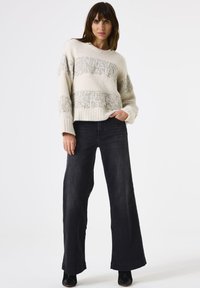 Eine Frau trägt einen cremefarbenen Strickpullover mit silbernen Streifen und weiten schwarzen Jeans, steht mit einer Hand in der Tasche vor einem weißen Hintergrund.