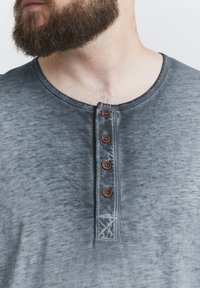 Šedé henley tričko vyrobené z měkké látky s texturovaným povrchem, se zaobleným výstřihem a čtyřmi hnědými knoflíky podél légy.