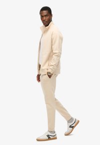 Chaqueta beige con cremallera y pantalones a juego de tela suave, con un diseño ajustado. Camisa blanca debajo y zapatillas deportivas blancas con acentos negros.