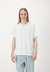 Balta medvilninė marškinėliai su tekstūruotu LEVI'S logotipu ant krūtinės, apvaliu kaklu ir laisvu siluetu. Suporuota su šviesiai mėlynomis džinsinėmis kelnėmis.