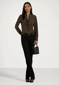 Mulher em pé, vestindo uma blusa castanha estampada, calças pretas, sapatos de salto pretos e segurando uma pequena mala preta.