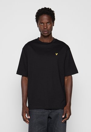 Mann mit kurzen lockigen Haaren, der ein schwarzes, übergroßes T-Shirt mit kleinem gelben Vogel-Logo auf der Brust und dunkle Jeans trägt.