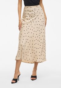 Bež svilen midi krilo s tekočim dizajnom, ki vsebuje črne pike in rahlo razširjen rob. Nosi se z črnimi čevlji s odprtim nosom.