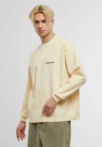 T-shirt en coton à manches longues de couleur jaune pâle avec un col rond, présentant un petit logo "ANOTHER COTTON" en impression foncée sur la poitrine.