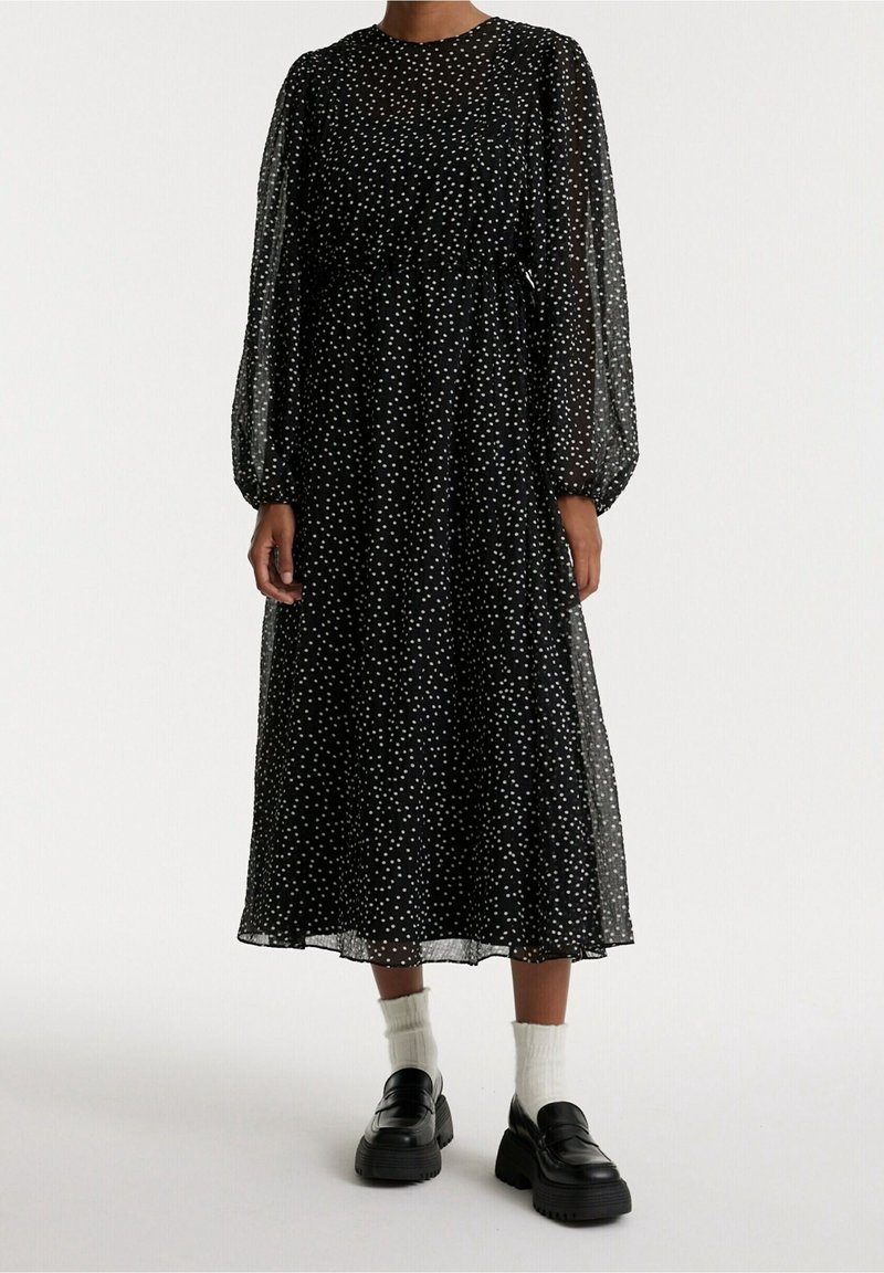 Schwarzes Polka-Dot-Kleid mit langen durchsichtigen Ärmeln, gesmokter Taille und knöchellangem Rock, kombiniert mit klobigen schwarzen Schuhen und weißen Socken.