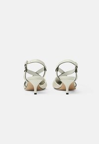 Sandalias de tacón de cuero blanco con tacón estrecho, diseño de tiras cruzadas y hebilla ajustable en el tobillo. Textura suave con un estilo minimalista.