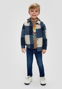 Blauw en crèmekleurig geruit overhemdjack, met knoopsluiting, met een bruine graphic t-shirt eronder, gecombineerd met donkere spijkerbroeken en witte sneakers.