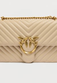 Μπεζ καπιτονέ δερμάτινη τσάντα χειρός με λουράκι αλυσίδα, διακοσμητικό χρυσό δαχτυλίδι και δύο διακοσμητικά πουλιά στο εμπρός καπάκι.