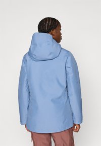 Ανοικτό γαλάζιο αδιάβροχο μπουφάν με κουκούλα, με λείο υφή και πλαϊνά φερμουάρ, σε συνδυασμό με ανοιχτό καφέ παντελόνι.