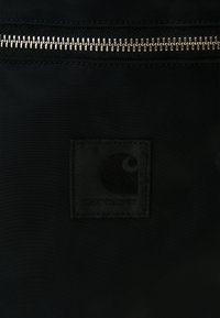 Bolsa de tela negra con un cierre plateado, que presenta un parche cuadrado de tela negra con el nombre de la marca "Carhartt."