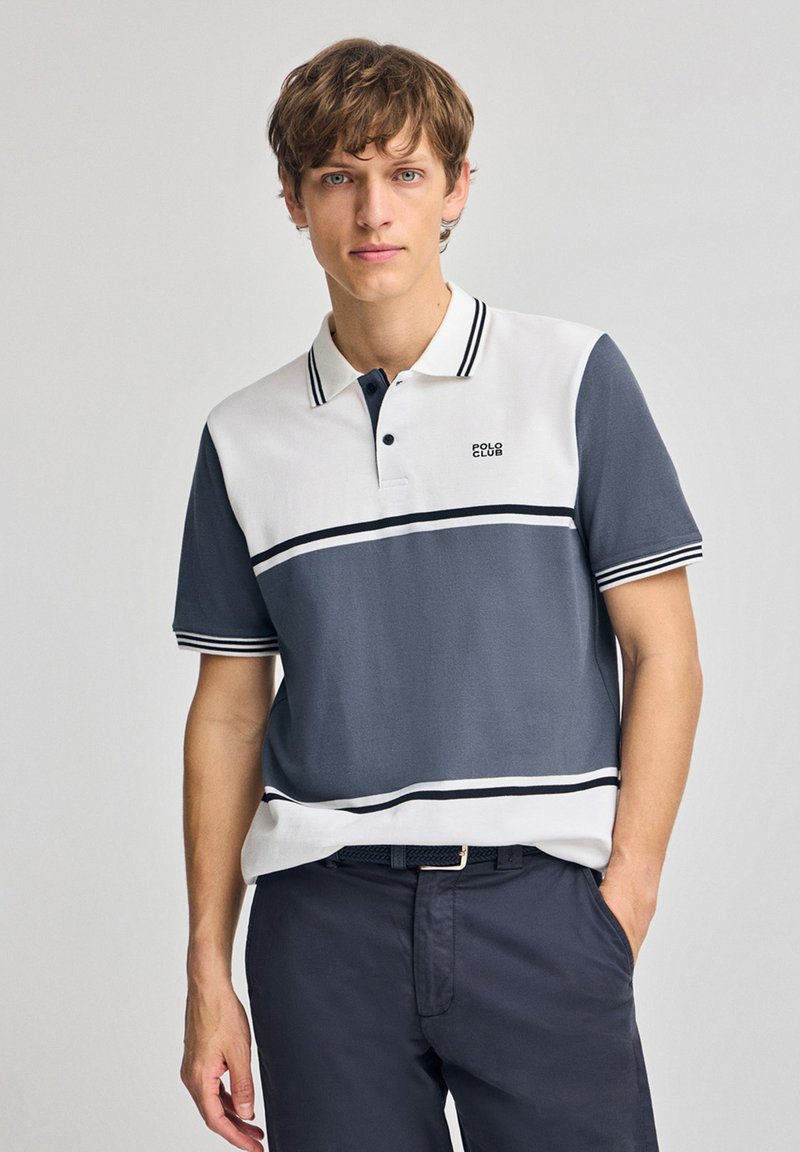 Poloshirt i vitt och marinblått, med horisontella ränder, krage och korta ärmar. Materialet verkar vara slätt med en avslappnad passform.