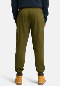 Olivgrüne Jogginghosen mit elastischem Bund und eng zulaufenden Knöcheln, aus weichem Stoff gefertigt. Getragen mit braunen Arbeitsstiefeln und einem dunklen Sweatshirt.