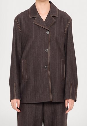 Brun korthåret jakke med hakket krave, tre knapper, forlommer og kontrastsyninger på ærmer og sømme, båret sammen med matchende bukser.