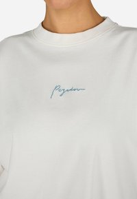 Fehér pulóver, kerek nyakkivágással, puha tapintással és világoskék hímzett "Pigador" logóval az elején. Kényelmes és lazán illeszkedő fazon.