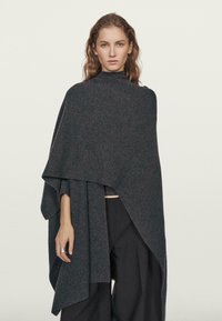 Žena s vlnitými vlasy v tmavě šedém asymetrickém pleteném ponču přes široké černé kalhoty, stojící na jednolitém světlém pozadí.