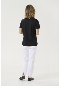 T-shirt noir à manches courtes, pantalon blanc avec des accents de nœuds à l'ourlet, chaussures plates noires, vue de dos sur un fond clair.