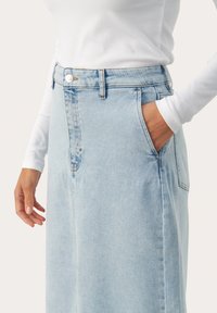 Jupe en denim bleu clair avec taille haute, poches avant et fermeture à boutons. Le tissu a une texture lisse et un léger délavage.