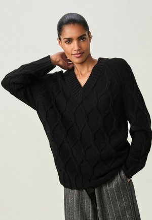 Sort strikket sweater med V-hals og diamantmønster, med løse ærmer og ribkant, parret med grå stribede bukser.