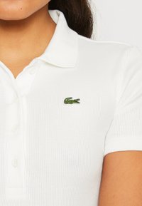Biely ryflový polo tričko s vyšitým zeleným logo krokodíla, s klasickým golierom a gombíkovou légou.