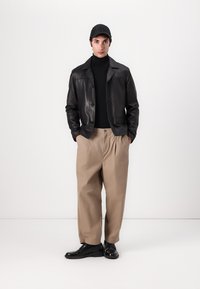 Jachetă din piele neagră peste un gât lung negru, pantaloni plisati bej și pantofi negri, completate cu o șapcă neagră. Texturi fine și croi ajustat.