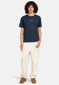 Navyblaues Baumwoll-T-Shirt mit kurzärmeligen Schlitzen und dem Text "Element". Kombiniert mit cremefarbenen, weit geschnittenen Hosen und braunen lässigen Schuhen.