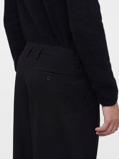 Person wearing black pants with belt loops and a back buttoned pocket, paired with a black long-sleeve top, hand relaxed by side.