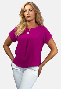 Fuksjowa bluzka z krótkimi, podwiniętymi rękawami, dekoltem w kształcie litery V i złotym guzikiem jako akcentem. Połączona z białymi spodniami z gładkiego materiału.