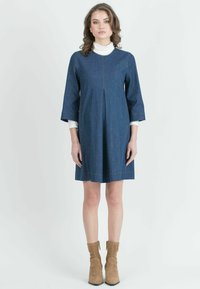 Denimimekko, jossa on pyöreä kaula-aukko, 3/4-hihat ja laskospintaa. Tummansininen kangas, jossa on kontrastiväriset oranssit saumat. Päällä valkoinen poolopaita.
