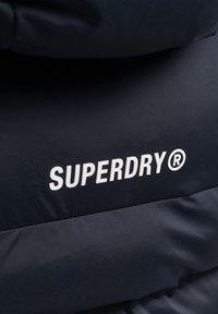 Nærbillede af sort jakke stof med hvid "SUPERDRY®" logo trykt på ryggen, der fremhæver mærket og teksturens detaljer.