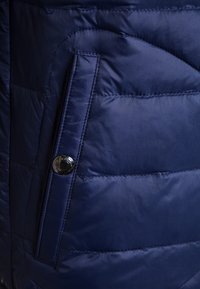 Gros plan d'une veste matelassée bleu marine avec des coutures horizontales et un détail de bouton pression sur le rabat d'une poche latérale.