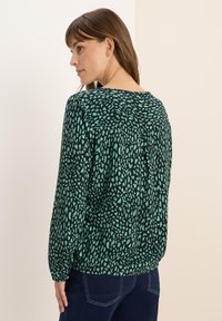 Grüne Bluse mit einem schwarzen abstrakten Muster, langen Ärmeln und rundem Ausschnitt. Hergestellt aus leichtem Stoff mit gerafftem Rückendetail.