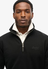 Pullover nero con zip e colletto alto, realizzato in tessuto testurizzato. Presenta una zip argentata e un sottile logo "SuperDry" sul petto.