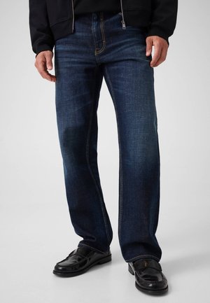 Mand iført mørkeblå jeans og sorte læderloafers står mod en ensfarvet lys baggrund.