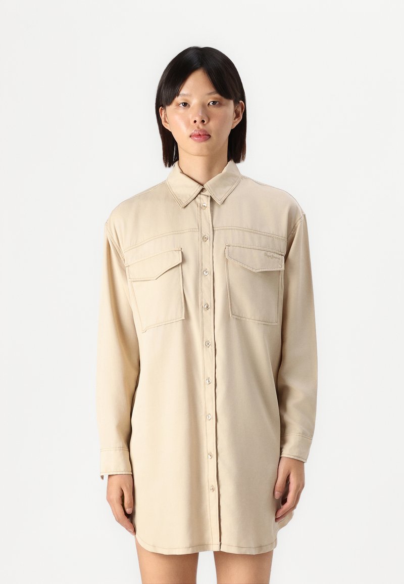 Abito camicia beige con maniche lunghe, caratterizzato da una chiusura con bottoni, due tasche sul petto e una vestibilità comoda, realizzato in un tessuto morbido con cuciture a contrasto.