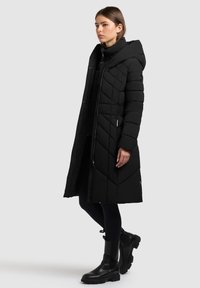 Zwarte gewatteerde pufferjas met een hoge kraag en een capuchon, voorzien van een rits en zijzakken. Gecombineerd met zwarte enkellaarsjes.
