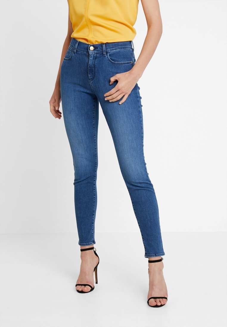 Jeans ajustados en denim azul medio con una cintura alta, un ligero efecto desgastado y estilo de cinco bolsillos, combinados con tacones negros de pulsera al tobillo.