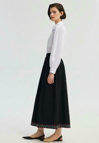 Fustă maxi neagră, cu broderie florală la tiv, asortată cu o cămașă albă cu nasturi. Modelul poartă sandale plate, cu vârful deschis.