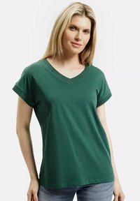 Blondynka w zielonym T-shircie z krótkimi rękawami i dekoltem w serek, w niebieskich dżinsach, stojąca na tle białej ściany.