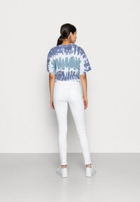 Sieviete ar tumšiem matiem astē, ģērbusies zilā un baltā krāsā raibā krekliņā, baltās augstās džinsās un baltās kedās, stāv pretī vienkāršai sienai.