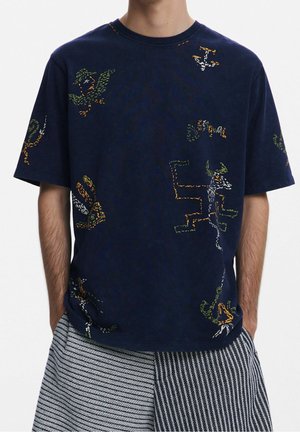 Homme portant un t-shirt bleu marine avec des motifs d’animaux colorés brodés et un short rayé noir et blanc, mains dans les poches.
