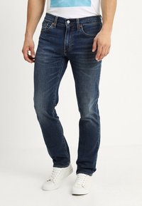 Ciemnoniebieskie dżinsy o prostym kroju, z dwiema przednimi kieszeniami i zapięciem na guzik. Noszone z białą koszulką i białymi trampkami.