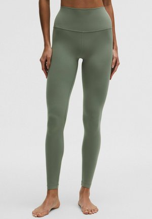 Kobieta ubrana w legginsy z wysokim stanem w oliwkowo-zielonym kolorze, stojąca boso na jednolitym jasnym tle.