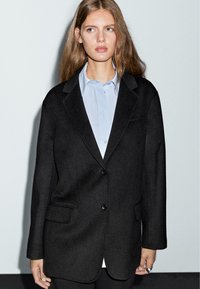 Černý vlněný blazer s klopami, jednoduchým zapínáním na jeden knoflík, dvěma kapsami a volným střihem, nošený přes světlomodrou košili.