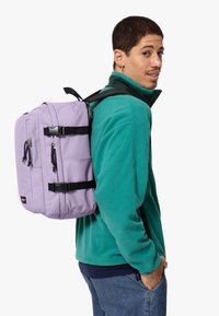 Mochila morada con correas negras ajustables, múltiples compartimentos con cremallera y hebillas. El modelo lleva una chaqueta de forro polar verde.