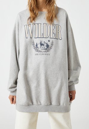 Personne portant un sweat-shirt gris oversize avec le texte "WILDER" et une illustration de cerf, montagnes et arbres au-dessus du texte "GREAT JOURNEY".